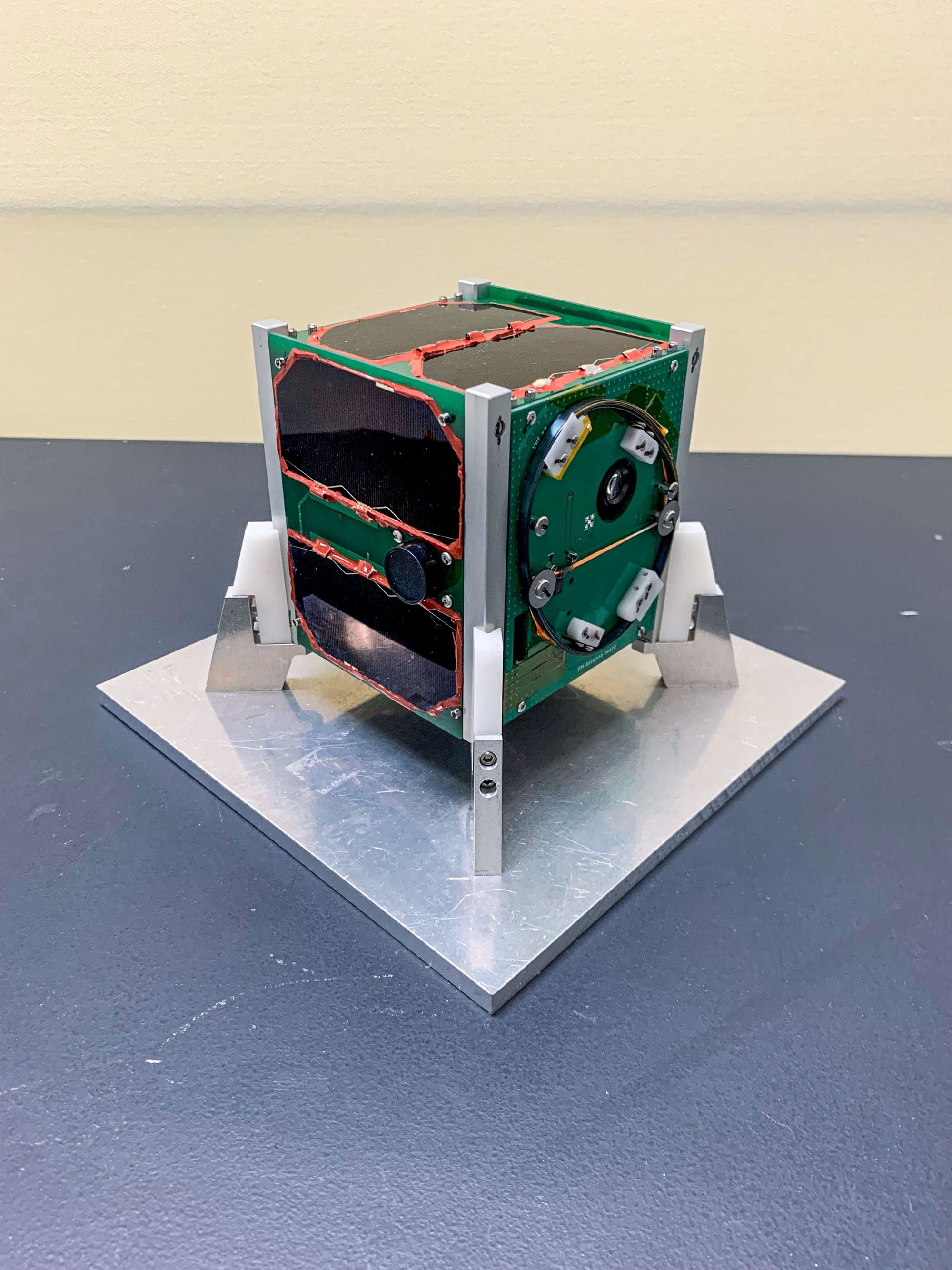 A one-unit CubeSat with solar panels and a camera aperture, standing on a bench