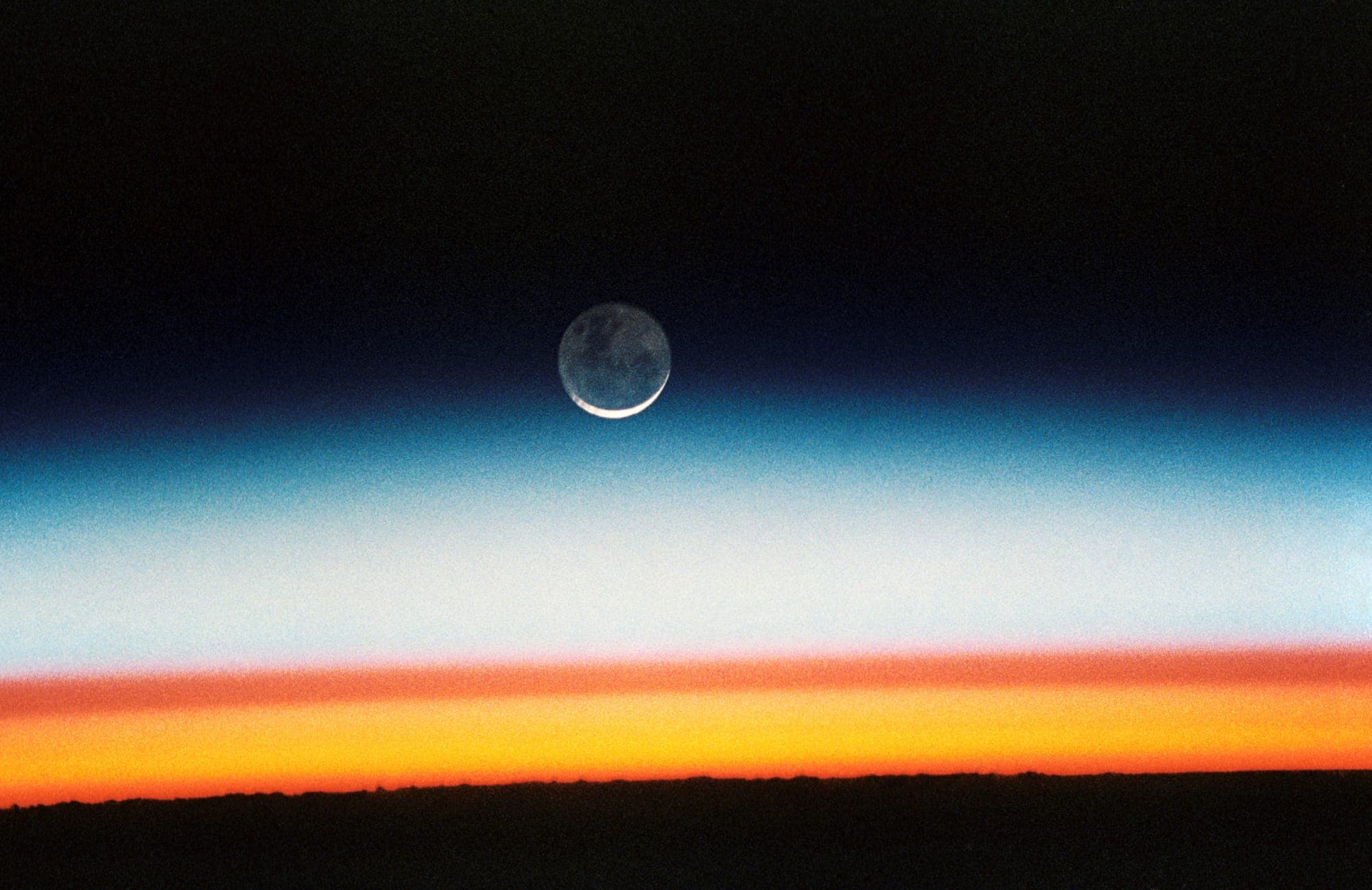 Sunrise on the curve of the Earth, with the crescent Moon above the layered atmosphere